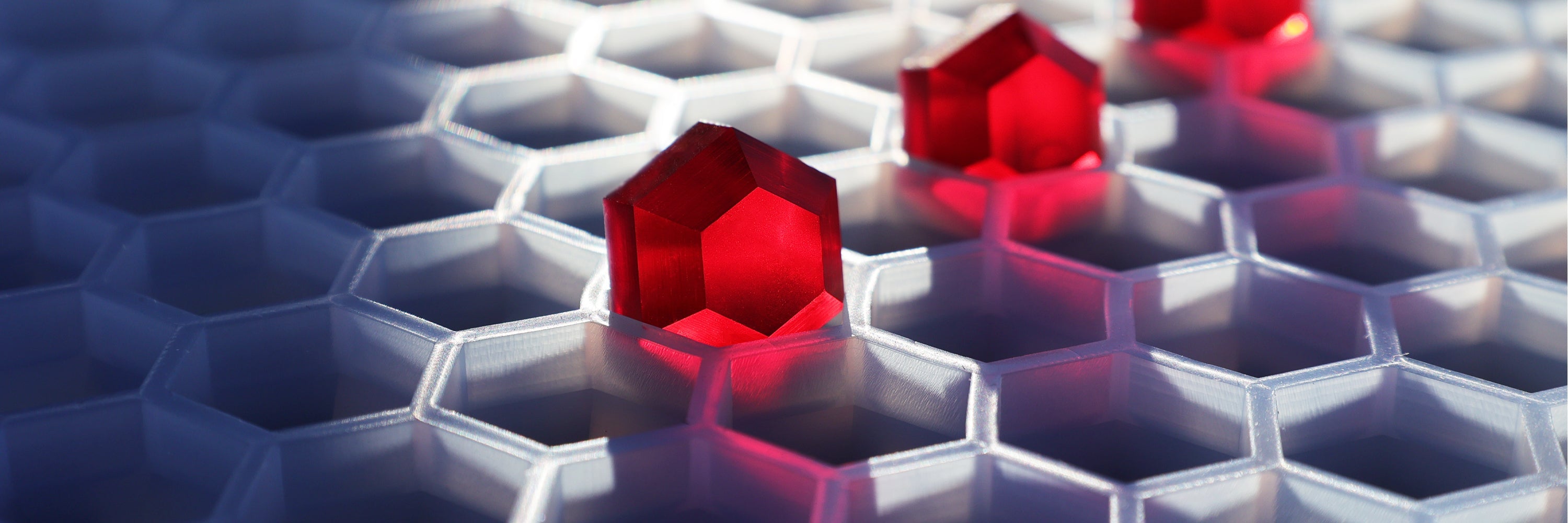 Red translucent house-shaped game pieces sit in a white hexagonal grid, illuminated by light and casting shadows.
