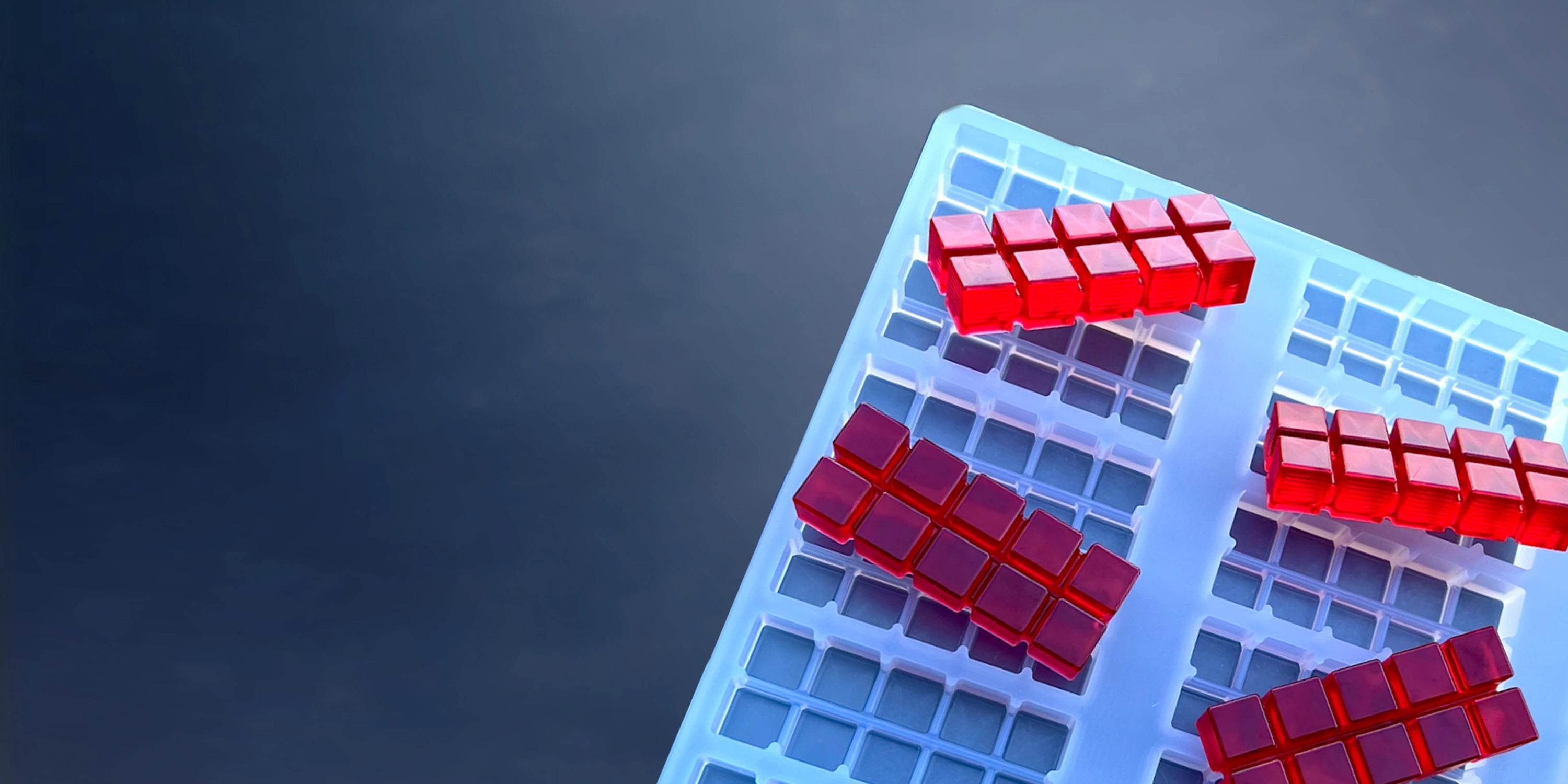A close-up of a blue ice cube tray holding several red gelatin cubes arranged in clusters, with a dark gradient background.