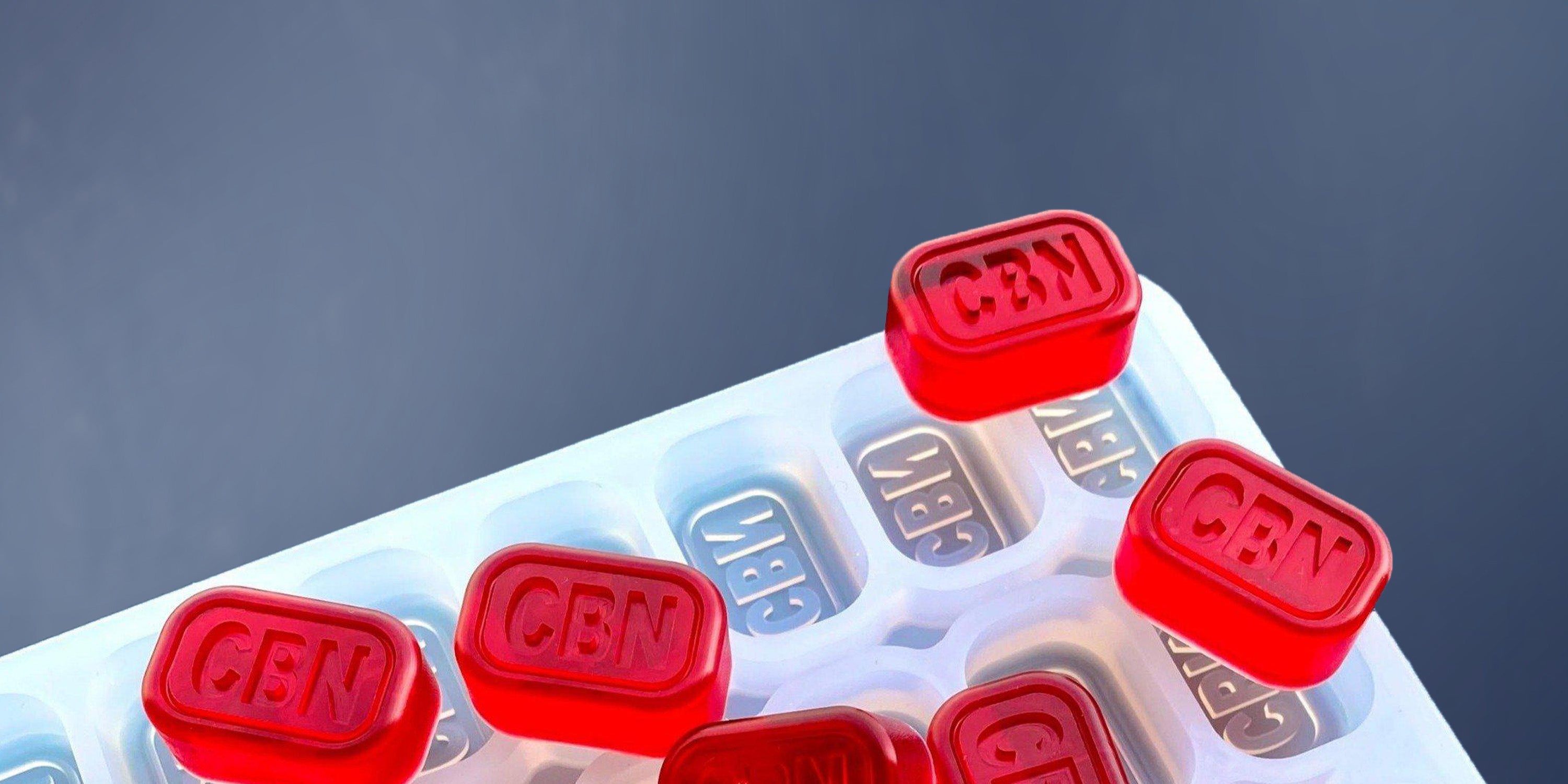 Red translucent gummies labeled "CBN" rest on and inside a white plastic tray with corresponding "CBN" labels, set against a plain blue background.