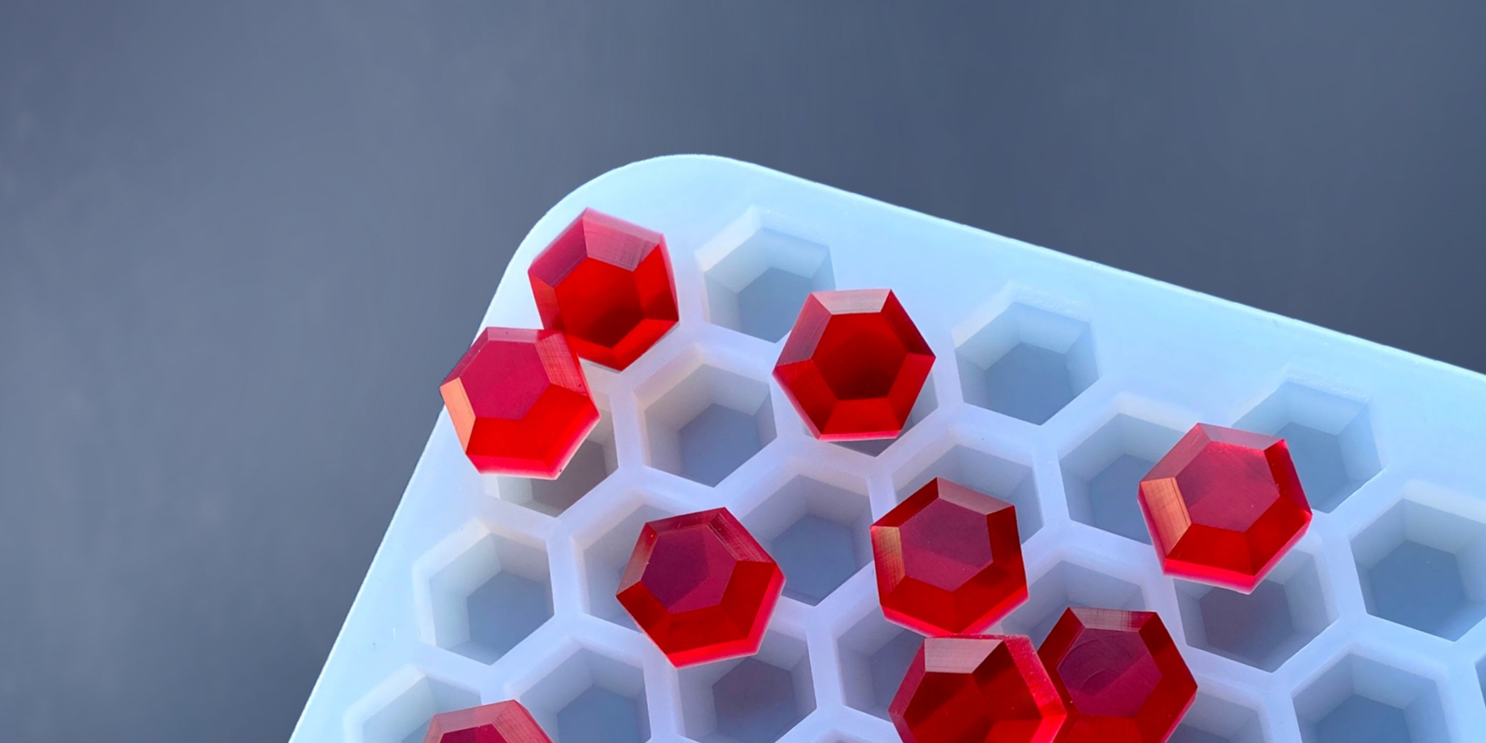 A white hexagonal ice tray contains several red hexagonal gelatin pieces, some compartments empty, against a gray background.