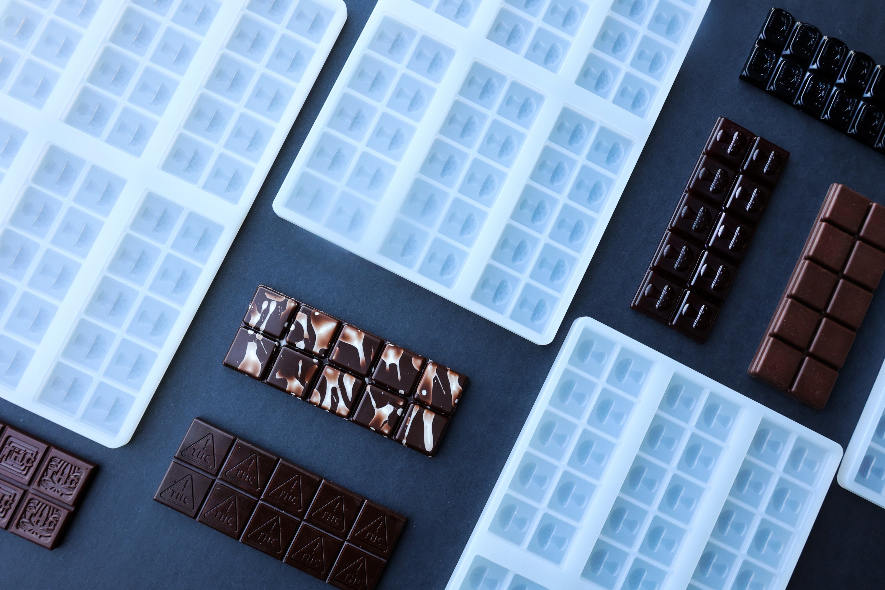 Assorted white silicone chocolate molds and various chocolate bars are arranged on a dark surface.