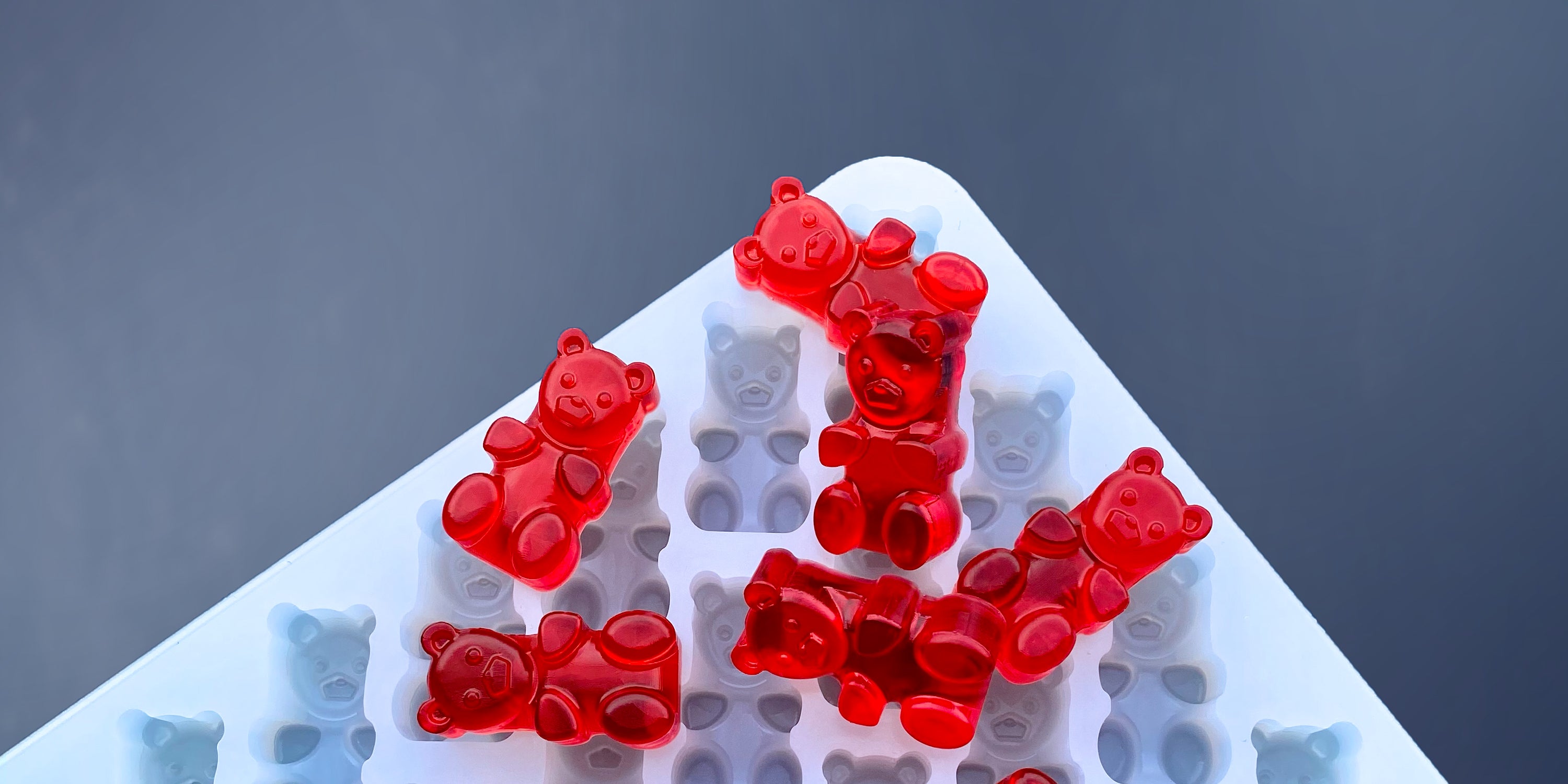 Red gummy bears are placed on top of a white silicone mold shaped like gummy bears, with a gray background.