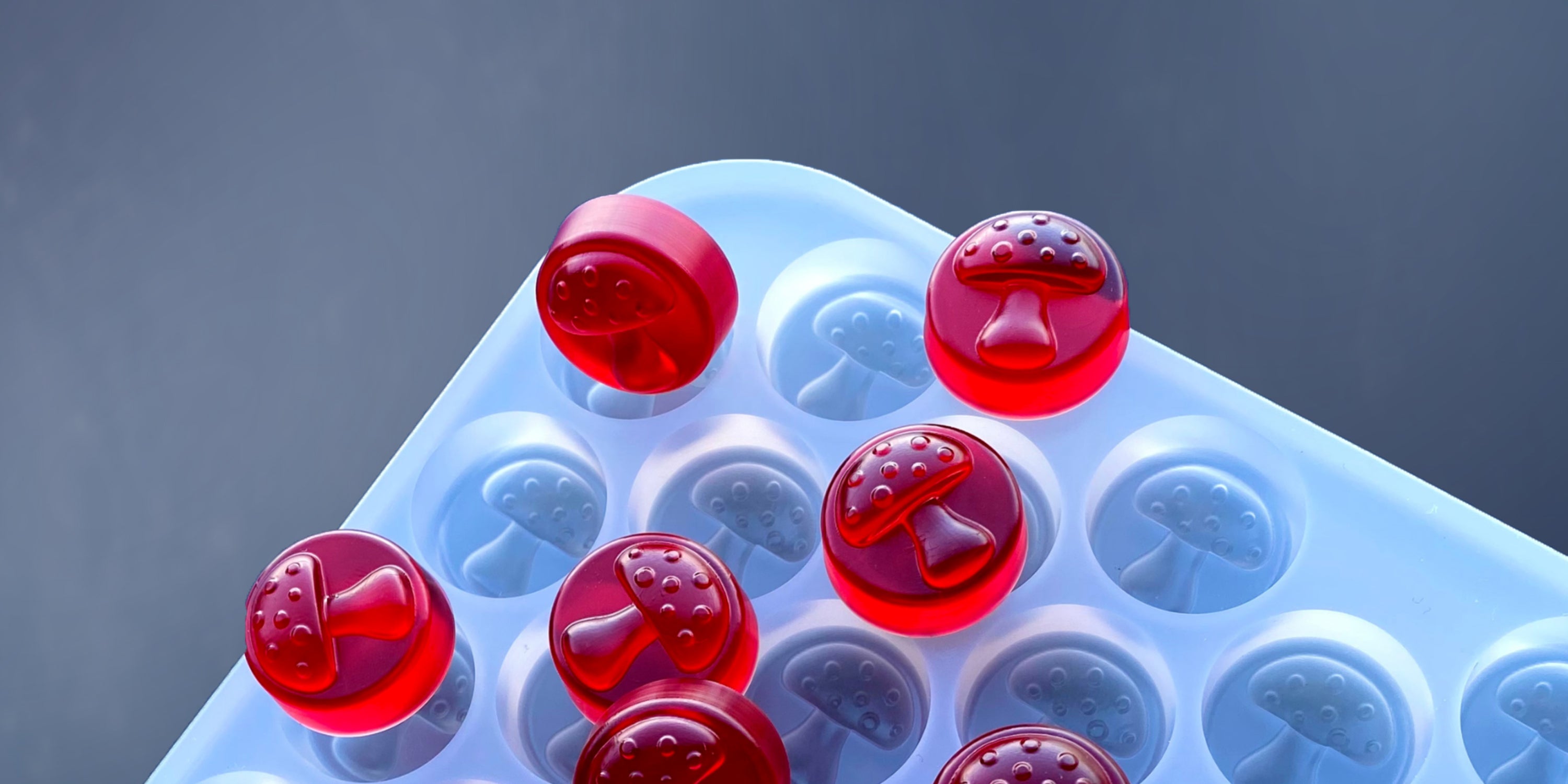 Red mushroom-shaped gummies on a light blue silicone mold with mushroom-shaped cavities, set against a plain background.