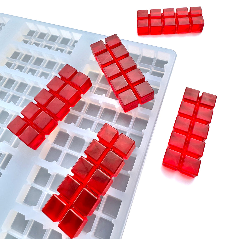 Several red, rectangular gelatin blocks with grid patterns are placed on and around a white Vector Molds 10 mL 3D Multidose Bar Gummy Mold - Universal Depositor, which features 60 matching rectangular cavities.