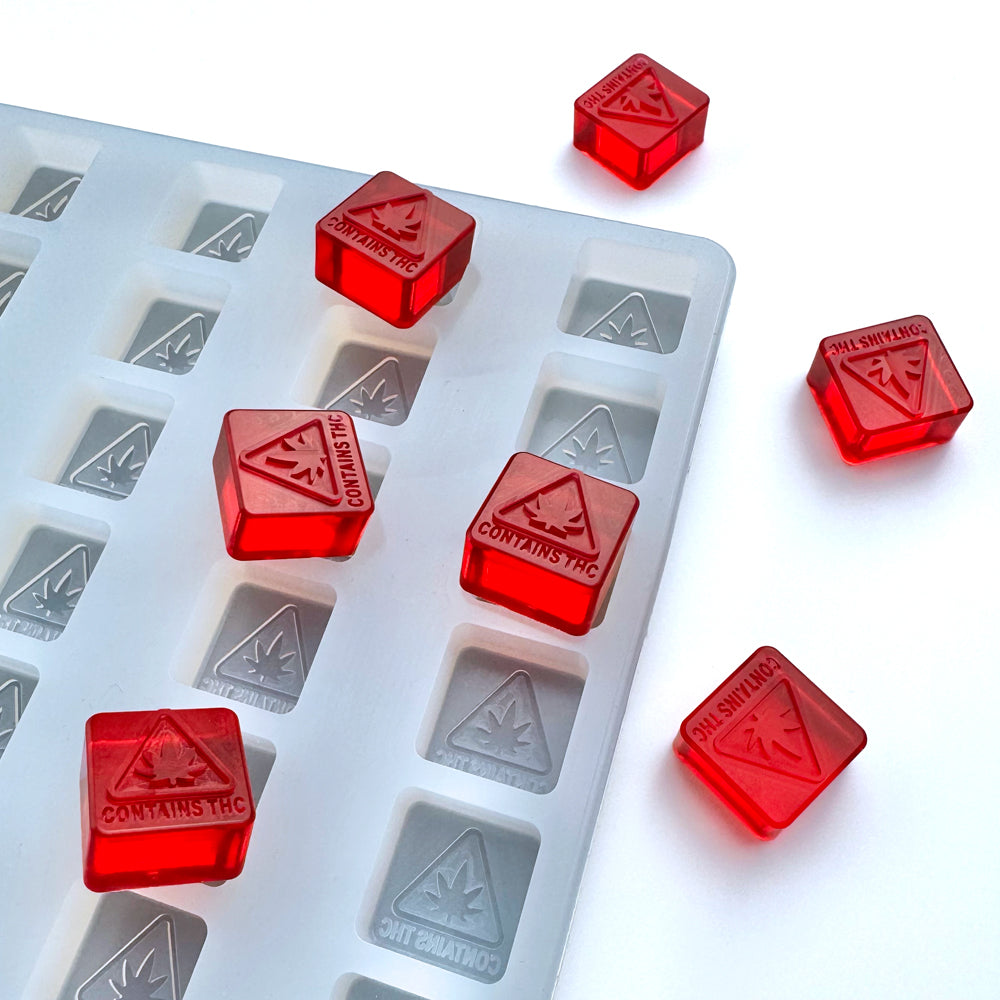 The 3 mL Gummy Mold - MA, ME, & RI THC Logo Mold by Vector Molds is shown with red, triangle-shaped gummies labeled "CONTAINS THC" placed on and around the professional food-grade silicone tray with 144 cavities.