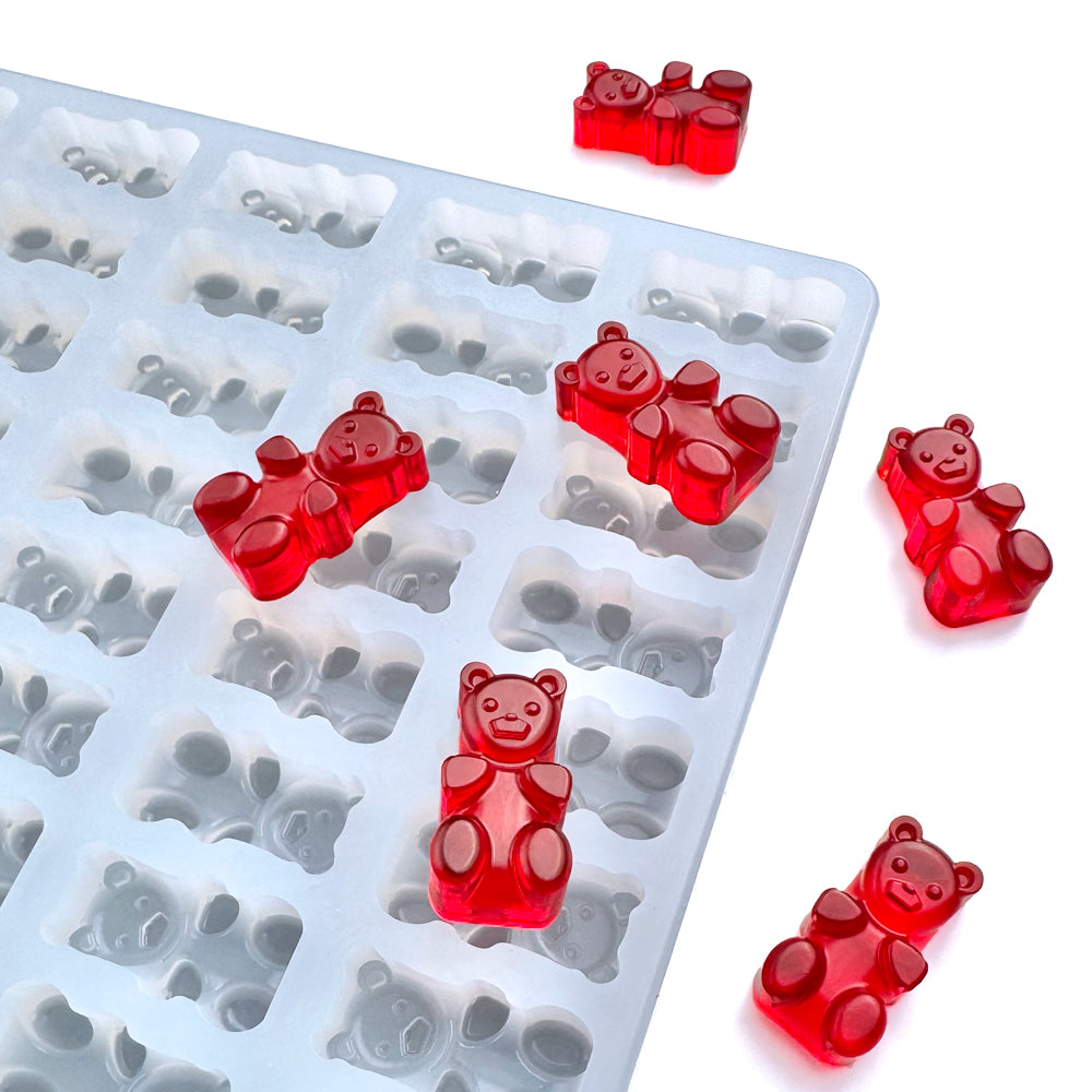 Red gummy bears rest on a white surface next to the Vector Molds 3 mL Gummy Bear Mold - Universal Depositor, featuring 176 cavities—perfect for making your own delicious sweets at home.
