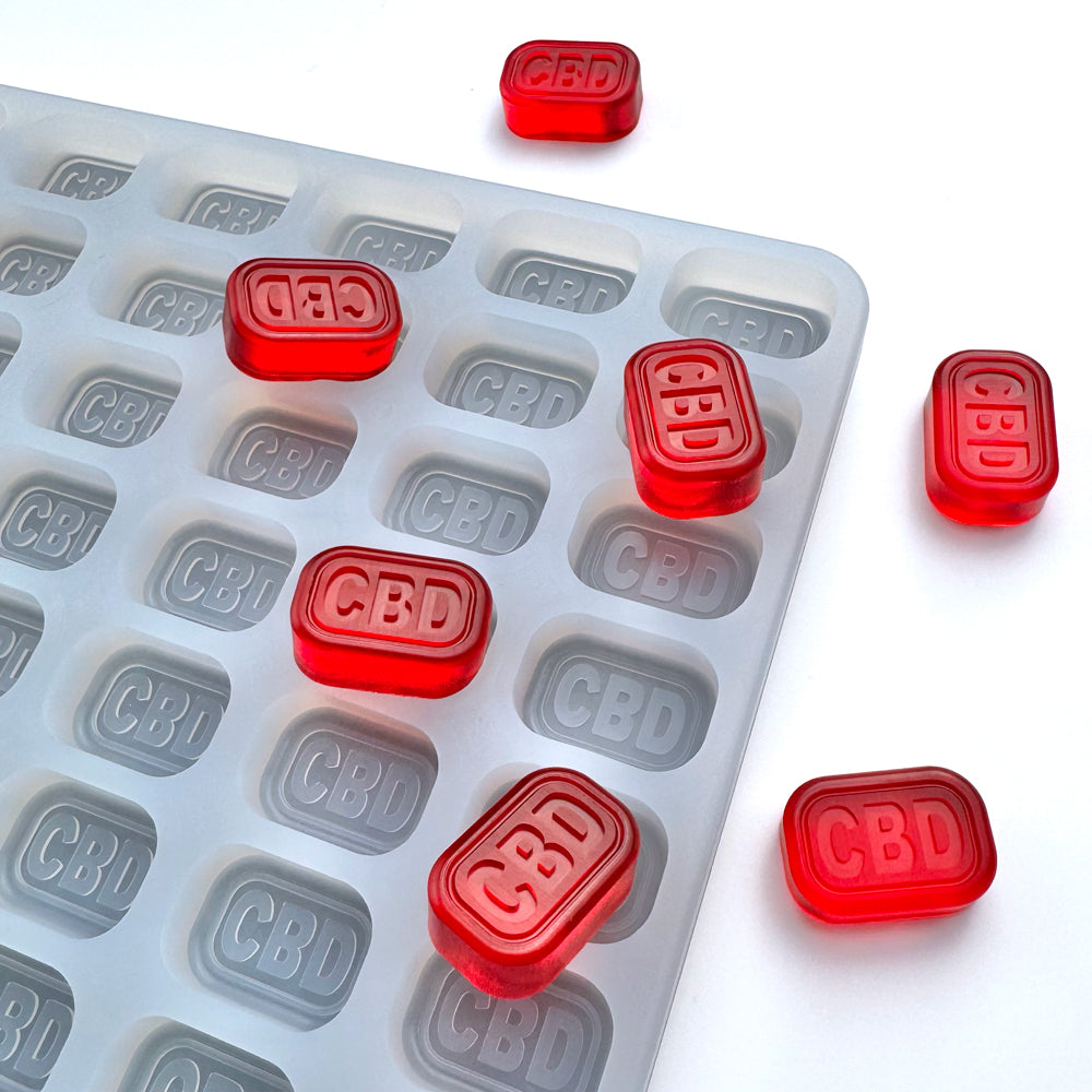 Red rectangular gummies labeled "CBD" sit on and around the Vector Molds 4 mL Brick Gummy Mold - Universal Depositor, a food-grade silicone tray with 144 cavities, each marked with the "CBD" logo.