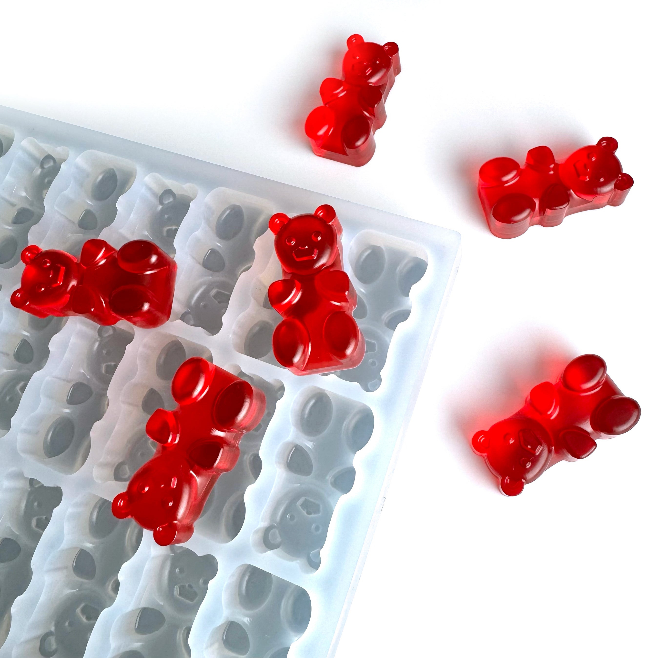Red gummy bears are placed on and around a white silicone mold with bear-shaped cavities, all set on a white background.
