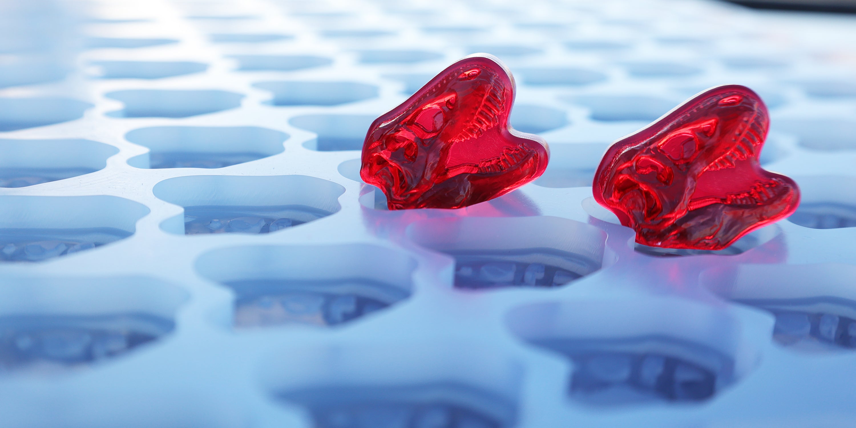Two red gummy candies shaped like dinosaur heads rest on a light blue silicone mold with multiple similar dinosaur head cavities.