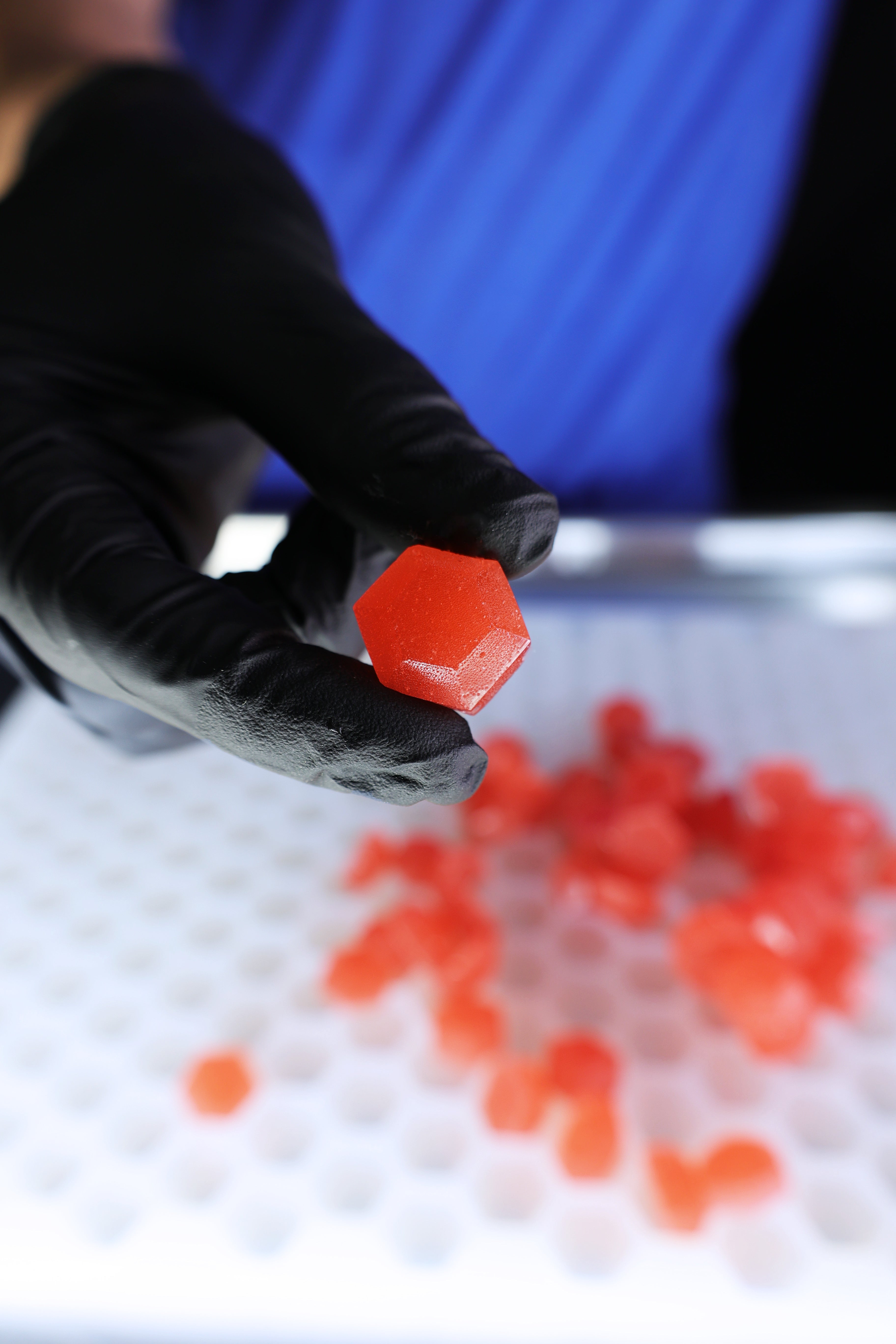 A gloved hand holds a red, hexagonal gummy candy above a tray with more gummies in the background.