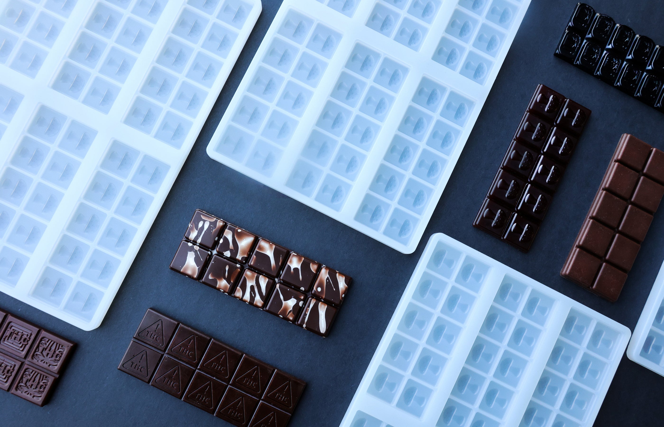 Assorted silicone chocolate bar molds and various finished chocolate bars arranged on a dark surface.