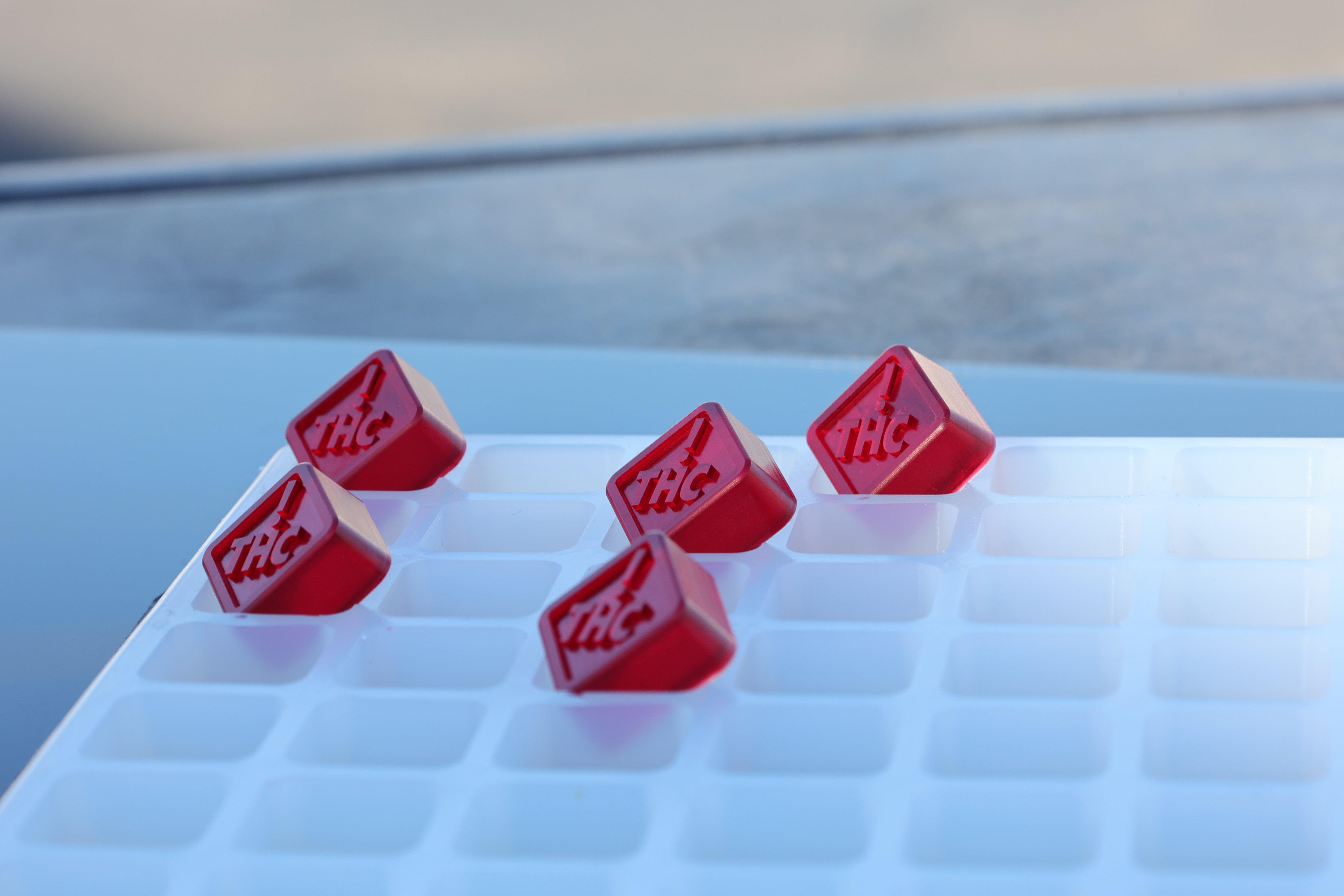 Five red THC-labeled gummies rest on a white tray with empty square compartments, ideal for use with the Vector Molds 2.5 mL Square Gummy Mold (CO, OH, NM & FL THC Logo), half sheet with 262 cavities—food-grade and BPA free.