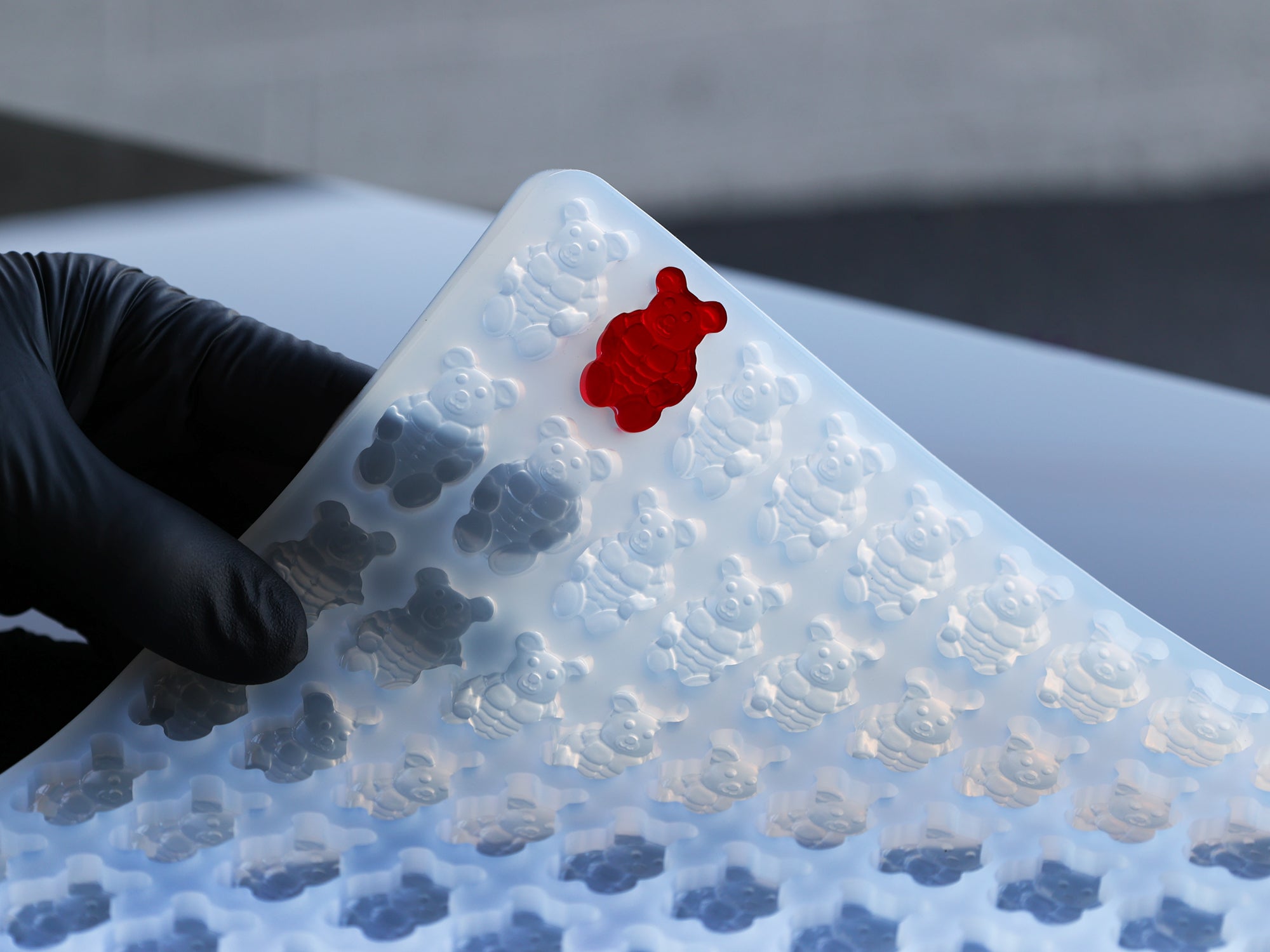 A gloved hand lifts a flexible silicone mold filled with bear-shaped cavities; one cavity contains a single red gummy bear.