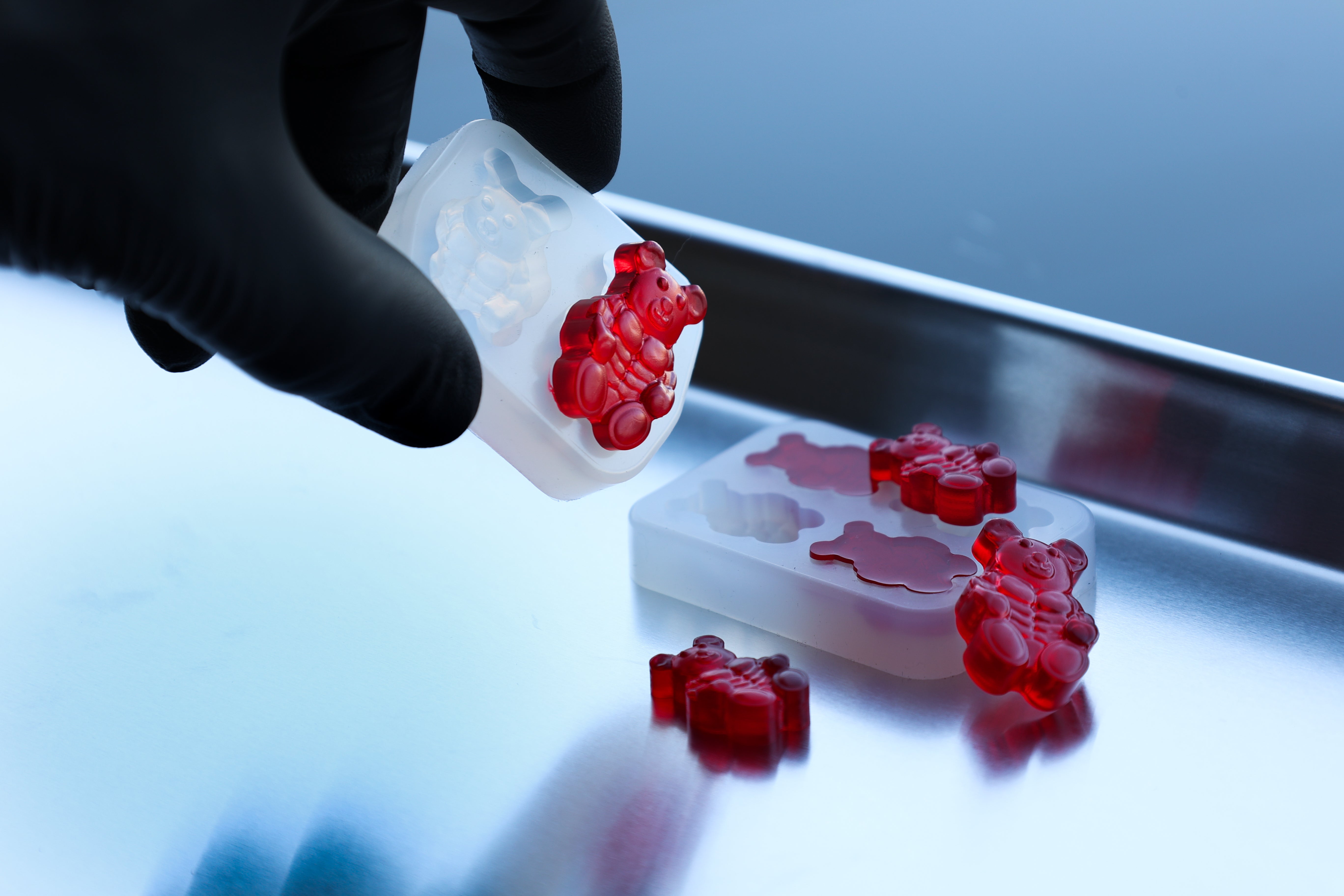 A gloved hand holds a silicone mold with red gummy bears, with more gummies and an empty mold section on a metal tray.