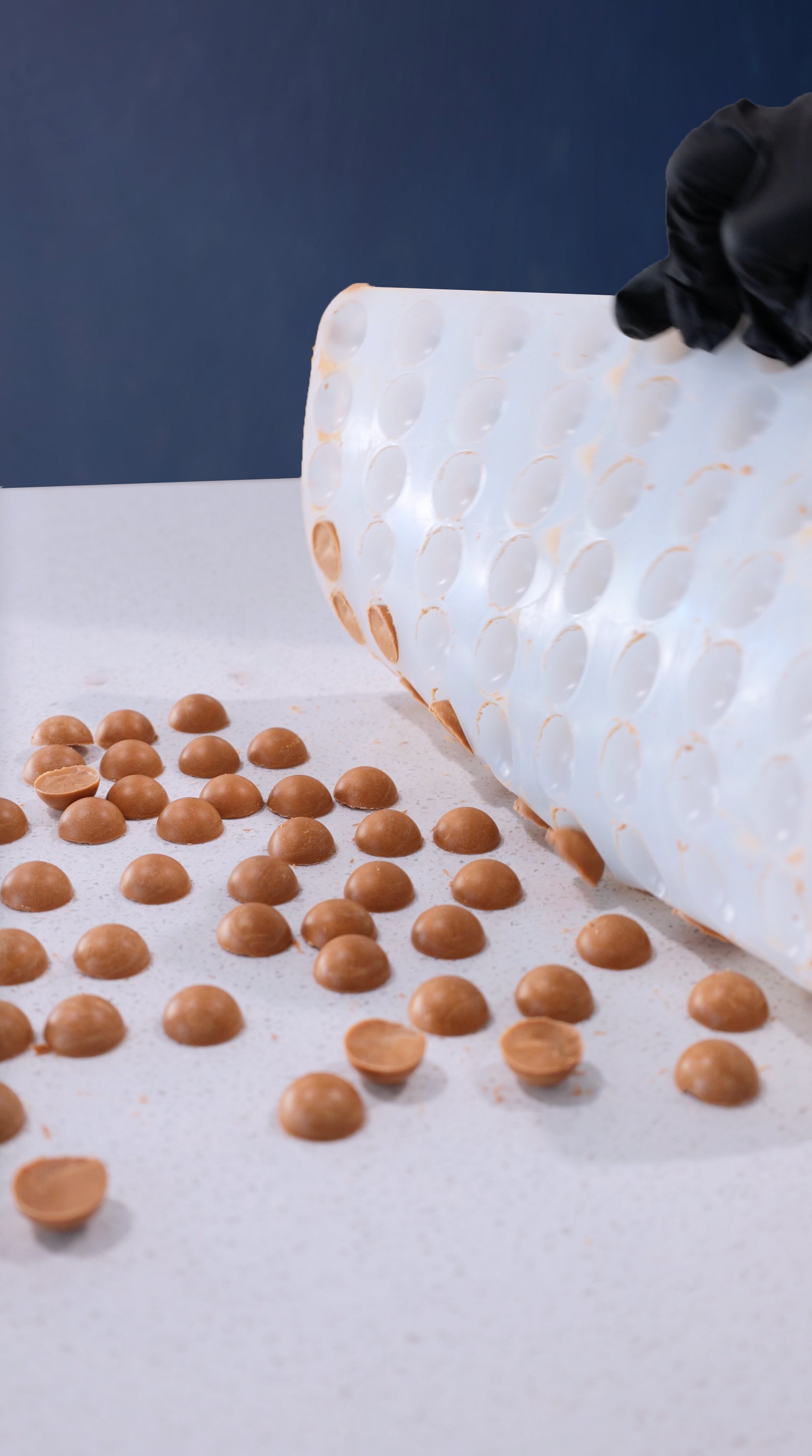 A gloved hand lifts a flexible mold, releasing small, round caramel candies onto a white surface.