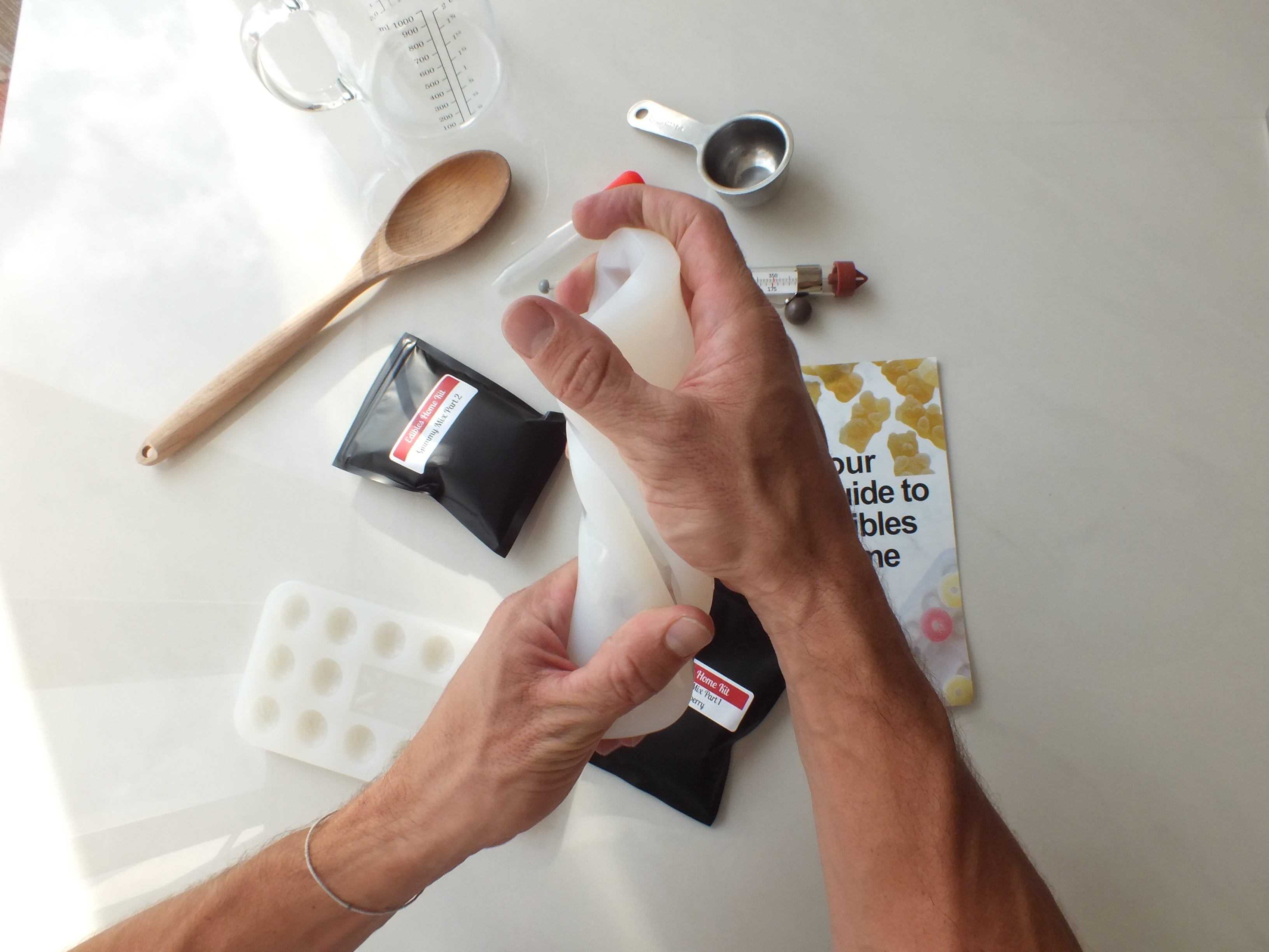 A person twists a white silicone mold over a table with black bags, a wooden spoon, a metal measuring cup, a makeup tube, a book, a measuring jug, and a white mold tray.
