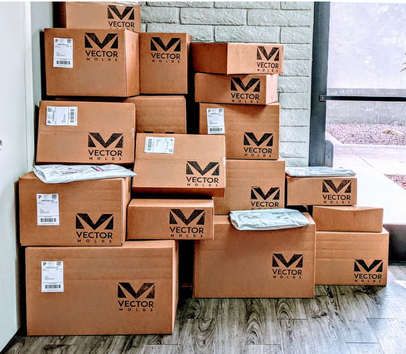 A stack of cardboard boxes labeled "Vector Molds" sits by a wall and window; some boxes have shipping labels and padded envelopes on top.