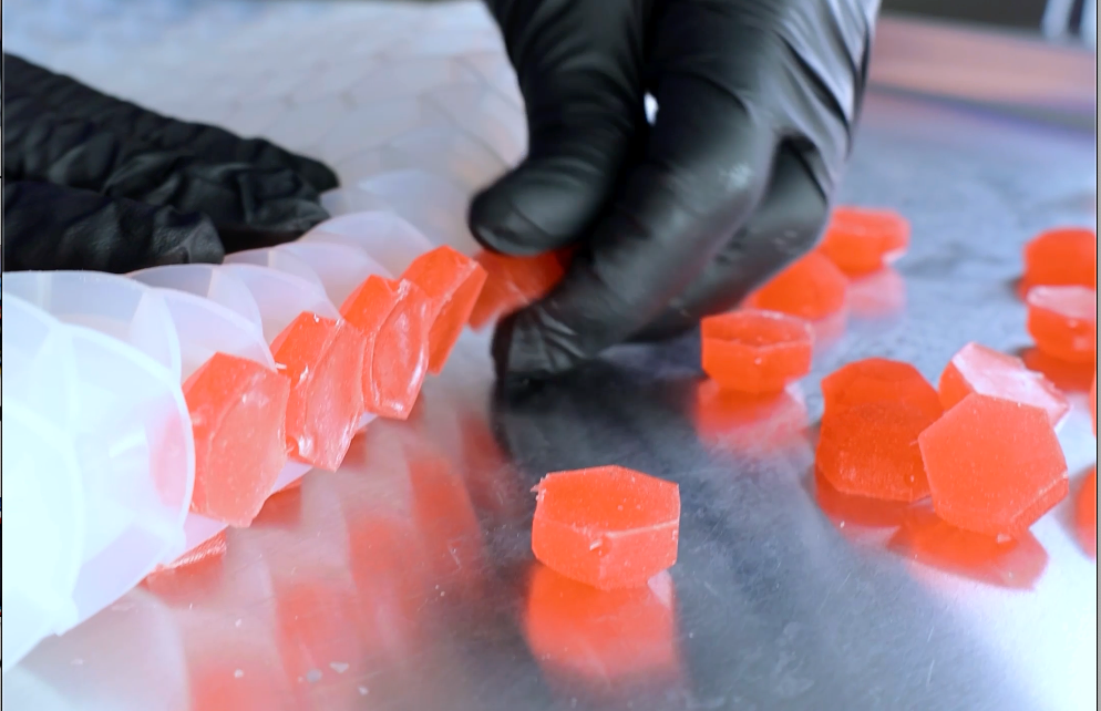 A person wearing black gloves arranges hexagonal red candies on a reflective metal surface.