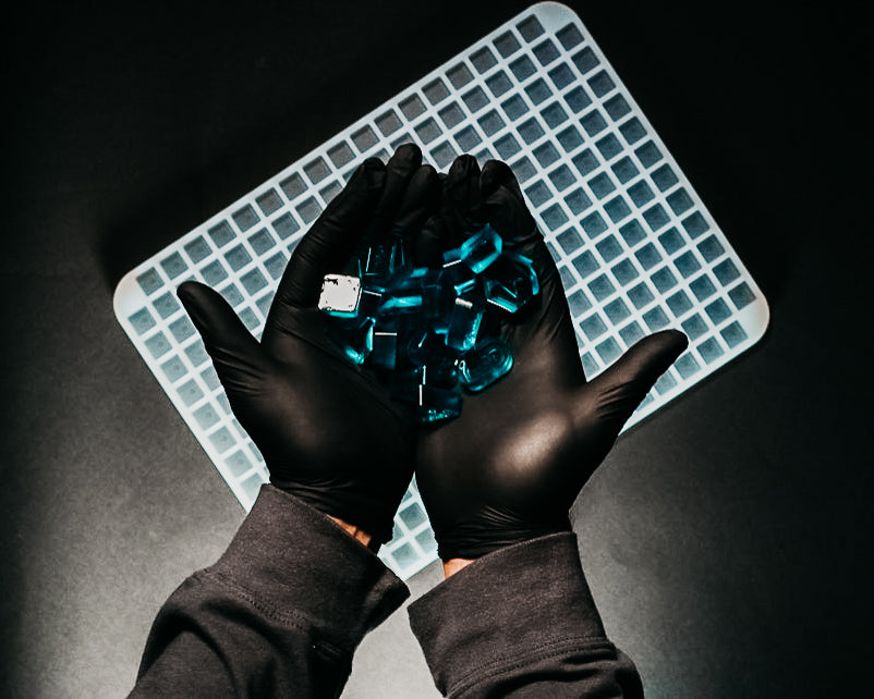 A pair of gloved hands holds blue transparent capsules over a white grid tray on a dark background.