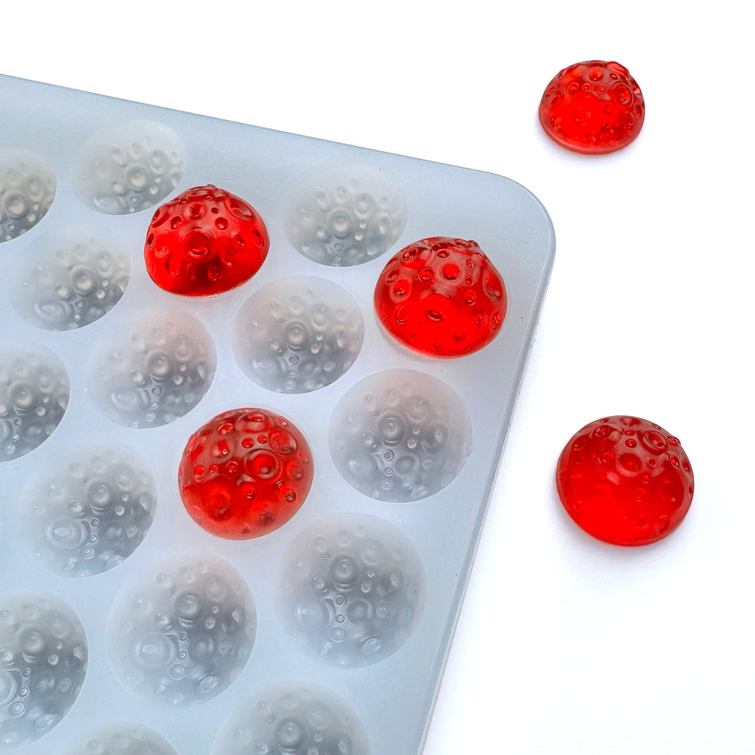 The Vector Molds 3.2 mL Moon Bon Bon Gummy Mold - Half Sheet with 195 cavities holds several red, dome-shaped gummies with a bubble texture, while two finished domes are displayed nearby on a white surface.