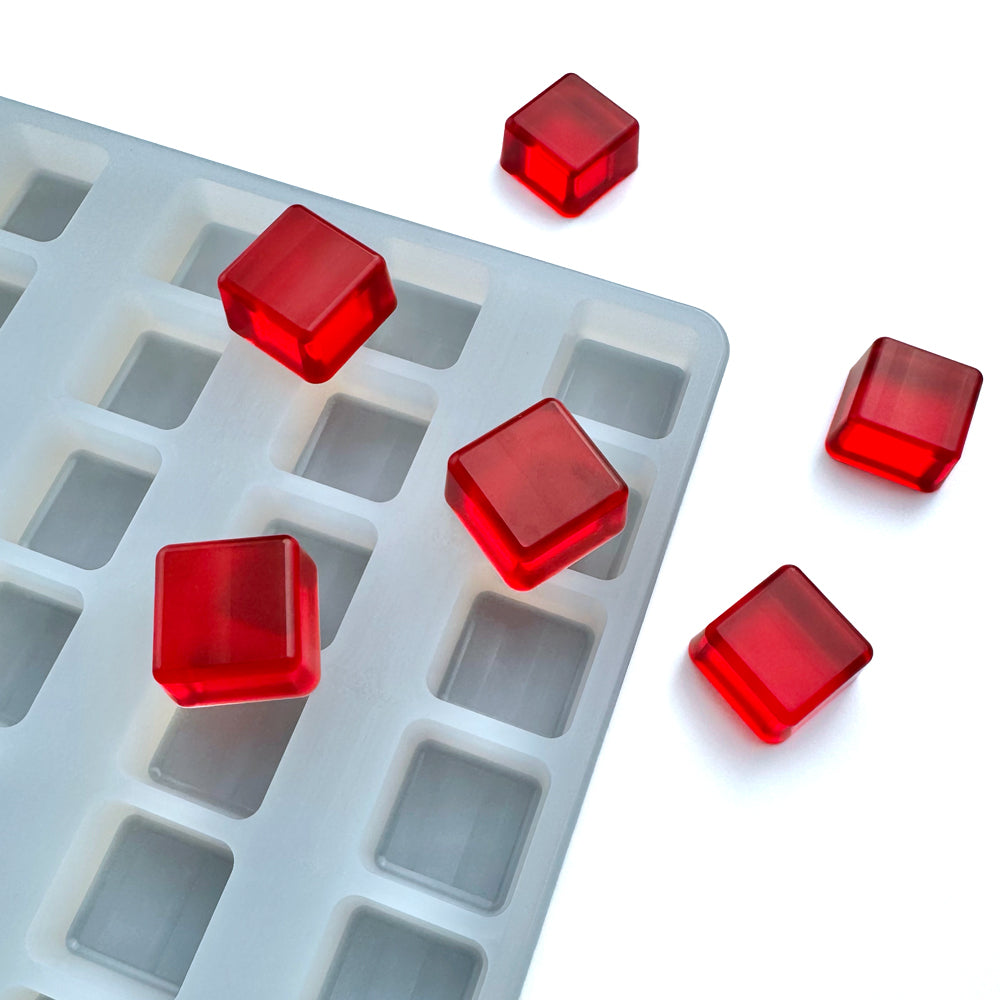 A close-up of the Vector Molds 4 mL Square Gummy Mold - Universal Depositor (136 Cavities) with bright red gelatin cubes inside and next to the transparent silicone tray on a white surface.